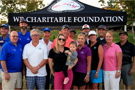 A group of people stand together in a park in front of a tent that reads “WE Charitable Foundation”